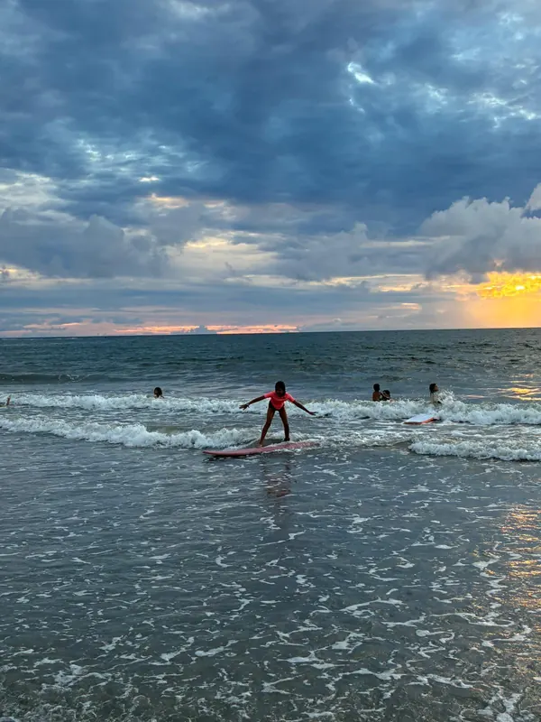 A Duckie rides a small evening wave towards the camera, arms out, under a sunset sky.